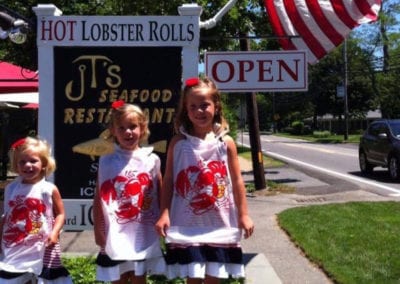3 girls standing with lobster roll aprons in front of JT's Seafood Restaurant sign